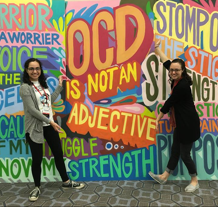 Co-Founders Morgan and Molly stand in front of a colorful painting framing the phrase, "OCD is not an adjective." Other words on the side include "strength" and "stomp out the stigma."
